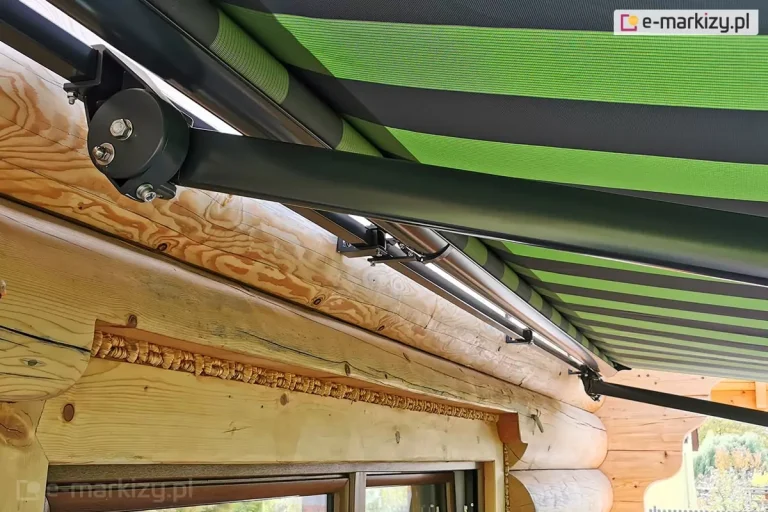 Szerokie ujęcie techniczne prezentujące sposób montażu oraz konstrukcję ramion markizy Adagio na budynku z bali drewnianych. Zdjęcie wykonane z dolnej perspektywy ukazuje dwa punkty podparcia markizy zamocowane do grubej, poziomej belki elewacyjnej. Konstrukcja wsporcza, rura nawojowa oraz ramiona przegubowe są wykonane z aluminium malowanego na matowy kolor ciemnoantracytowy. Po lewej stronie widoczny jest masywny uchwyt boczny z okrągłą maskownicą mechanizmu regulacji kąta nachylenia, z którego wychodzi długie, proste ramię markizy Adagio skierowane w prawą stronę. Wzdłuż rury nawojowej zamontowane są dodatkowe wsporniki stabilizujące cały system. Poszycie markizy Adagio wykonane jest z tkaniny w szerokie, poziome pasy w kolorach soczystej zieleni, ciemnej zieleni oraz czerni. Materiał jest w pełni rozwinięty i napięty, zasłaniając górną część kadru. Poniżej linii montażu markizy znajduje się fragment okna w brązowej ramie, a szczeliny między balami wypełnione są dekoracyjnym mszeniem z warkocza drzewnego. Naturalna struktura jasnego drewna z widocznymi słojami i sękami stanowi tło dla nowoczesnych, metalowych elementów markizy Adagio.