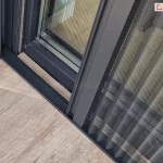 Zbliżenie prowadnicy dolnej moskitiery, w której widoczne są sznurki odpowiadające za rozwijanie i składanie plisowanej siatki. Próg w kolorze konstrukcji moskitiery zamocowany został do podłogi tarasu wyłożonego drewnopodobnymi płytkami w lekkim oddaleniu od ramy okna z uwagi na występujące przy nim zintegrowane rolety zewnętrzne.