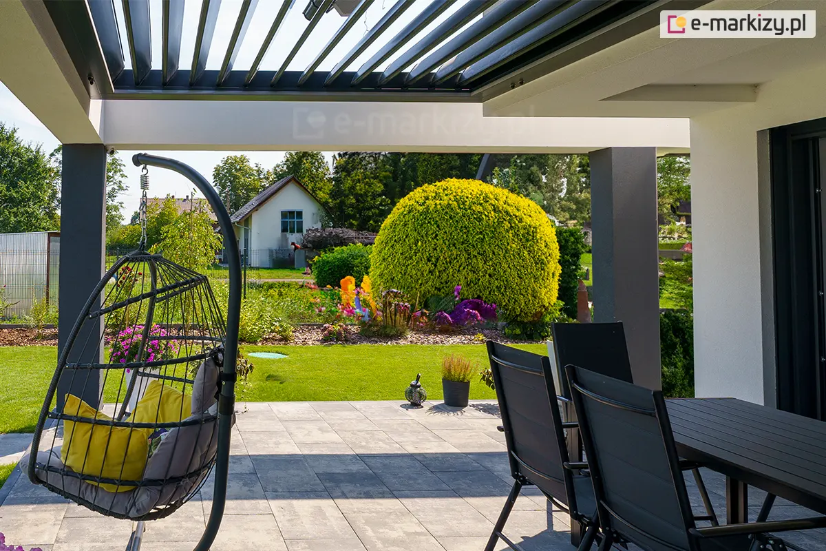 widok z wnętrza nowoczesnej pergoli SB400R na ogród, skupiając się na funkcjonalności ruchomych lameli dachowych. Aluminiowe lamele w kolorze antracytowym są ustawione w pozycji uchylonej pod kątem około 45 stopni, co pozwala na jednoczesną wentylację przestrzeni oraz naturalne doświetlenie tarasu przy zachowaniu ochrony przed bezpośrednim słońcem. Konstrukcja dachu opiera się na białych, szerokich belkach poprzecznych i ciemnoszarych słupach nośnych. Pod zadaszeniem znajduje się strefa wypoczynkowa z nowoczesnymi meblami: czarnym stołem z krzesłami oraz wiszącym fotelem typu "kokon" z szarymi poduszkami, zamontowanym na stabilnym stelażu. Posadzka tarasu została wykonana z dużych, jasnoszarych płyt betonowych. W tle rozpościera się widok na zadbany trawnik, formowane krzewy ozdobne o intensywnie żółtych i zielonych barwach oraz fragmenty sąsiedniej zabudowy pod pogodnym, jasnym niebem.