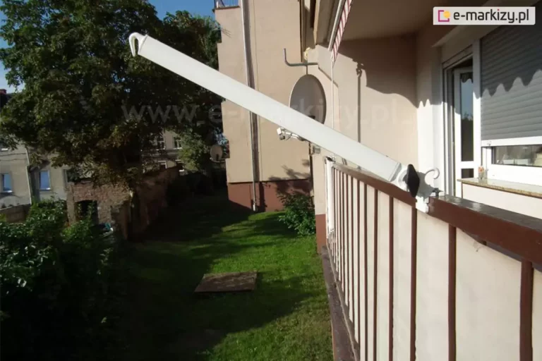 Zdjęcie przedstawia widok z boku na rozłożone ramię, w jakie wyposażona jest markiza balkonowo-poręczowa, zamontowana na balustradzie tarasu. Białe, aluminiowe ramię o prostokątnym przekroju jest uniesione pod kątem, skierowane na zewnątrz balkonu, a jego koniec wieńczy biały haczyk służący do zaczepienia tkaniny markizy. Konstrukcja jest przymocowana do brązowej, metalowej poręczy za pomocą białego uchwytu klamrowego, wyposażonego w czarną śrubę dociskową z ergonomicznym uchwytem. Wzdłuż barierki rozciągnięta jest beżowa osłona balkonowa, a pionowe szczeble balustrady są wyraźnie zaznaczone. W tle widoczna jest elewacja budynku w kolorze piaskowym z zainstalowaną białą anteną satelitarną oraz fragment okna z szarymi roletami zewnętrznymi. Na ścianie budynku, nad oknem, rzucany jest charakterystyczny ząbkowany cień pochodzący od falbany markizy. Perspektywa zdjęcia obejmuje również widok na podwórze z zieloną trawą, drzewem o gęstym liściu oraz fragmentem ceglanego muru w oddali. Całość oświetlona jest mocnym, naturalnym światłem słonecznym, podkreślającym detale techniczne mechanizmu pozycjonującego markizę.