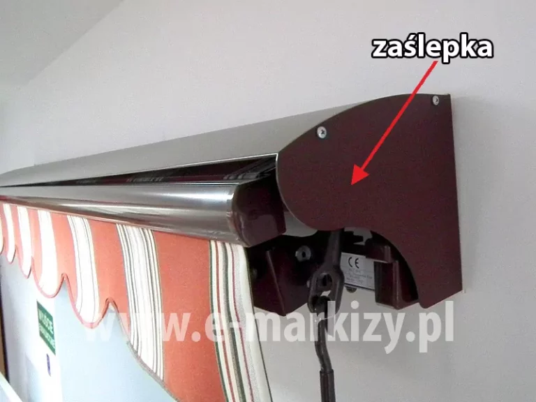 Markiza tarasowa z brązową konstrukcją i tkaniną falbany w czerwono-biało-zielone pasy o falowanym wykroju oraz widoczną przekładnią do napędu ręcznego, nad którą zamocowano uniwersalny daszek do markiz na białej ścianie. Po prawej stronie czerwona strzałka wskazująca boczek daszka, nad nią biały napis "zaślepka"