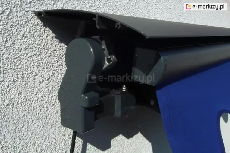 Lewy bok markizy bez kasety Aluprof w kolorze srebrzysty grafit z falbaną falowaną w kolorze chabrowym z widocznym nad belką przednią uniwersalnym daszkiem do markiz bez zaślepki bocznej, całość zainstalowana na ścianie o białym tynku strukturalnym