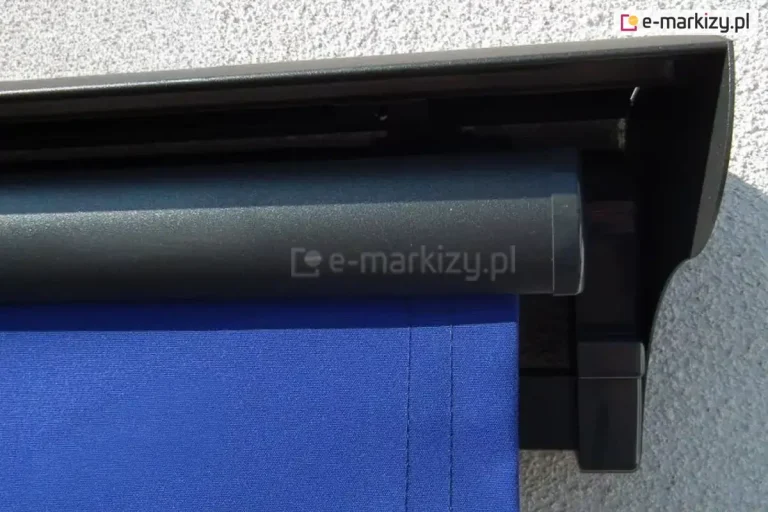 Prawy bok markizy tarasowej w kolorze srebrzysty grafit z chabrowym poszyciem oraz zamontowanym nad osłoną uniwersalnym daszkiem do markiz w kolorze konstrukcji markizy, całość zainstalowana na ścianie o białym tynku strukturalnym