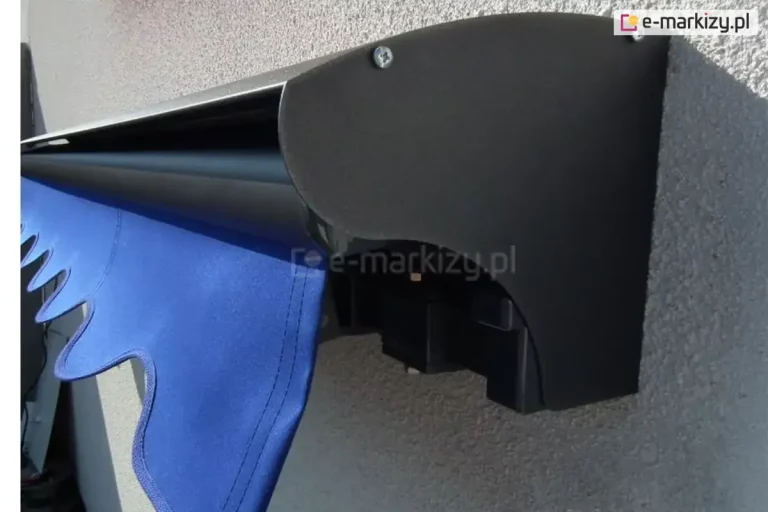 Markiza tarasowa bez kasety Aluprof z dodatkowym daszkiem do markiz chroniącym tkaninę poszycia przed warunkami atmosferycznymi, całość o konstrukcji w kolorze srebrzysty grafit, markiza z falbaną w odcieniu niebieskim o falowanym wzorze, zawieszona na ścianie o białym tynku strukturalnym widoczna od prawego boku
