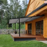 Lekka pergola tarasowa przyścienna Vera Sola o minimalistycznej konstrukcji aluminiowej w kolorze czarnym, zamontowana do elewacji klasycznego domu z bali drewnianych. Zadaszenie stanowi jednolite, gładkie poszycie tkaninowe w kolorze szarym, które jest całkowicie rozłożone pod skosem i wsparte na dwóch smukłych, czarnych profilach pionowych przymocowanych do krawędzi tarasu. Taras wykonany jest z brązowych desek kompozytowych i osadzony na ciemnej podmurówce, a prowadzą na niego niskie, drewniane schodki. Na tarasie, obok słupa konstrukcyjnego, ustawiono dwie donice z kwitnącymi roślinami. W tle widoczny jest wysoki, drewniany płot o nieregularnym wykończeniu oraz gęsty las iglasty pod lekko zachmurzonym niebem. Zdjęcie wykonane w świetle dziennym, prezentuje estetyczny kontrast między nowoczesnym systemem zadaszenia a tradycyjną architekturą drewnianą budynku.