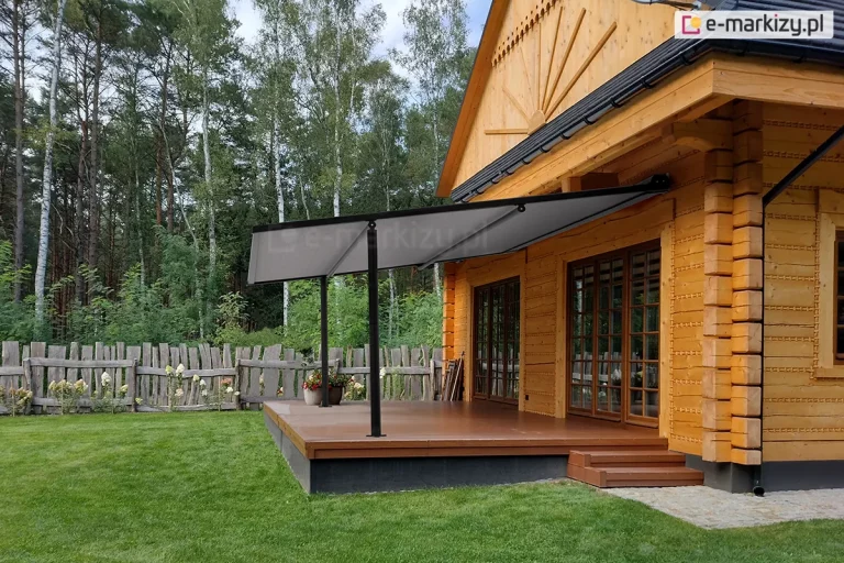 Lekka pergola tarasowa przyścienna Vera Sola o minimalistycznej konstrukcji aluminiowej w kolorze czarnym, zamontowana do elewacji klasycznego domu z bali drewnianych. Zadaszenie stanowi jednolite, gładkie poszycie tkaninowe w kolorze szarym, które jest całkowicie rozłożone pod skosem i wsparte na dwóch smukłych, czarnych profilach pionowych przymocowanych do krawędzi tarasu. Taras wykonany jest z brązowych desek kompozytowych i osadzony na ciemnej podmurówce, a prowadzą na niego niskie, drewniane schodki. Na tarasie, obok słupa konstrukcyjnego, ustawiono dwie donice z kwitnącymi roślinami. W tle widoczny jest wysoki, drewniany płot o nieregularnym wykończeniu oraz gęsty las iglasty pod lekko zachmurzonym niebem. Zdjęcie wykonane w świetle dziennym, prezentuje estetyczny kontrast między nowoczesnym systemem zadaszenia a tradycyjną architekturą drewnianą budynku.