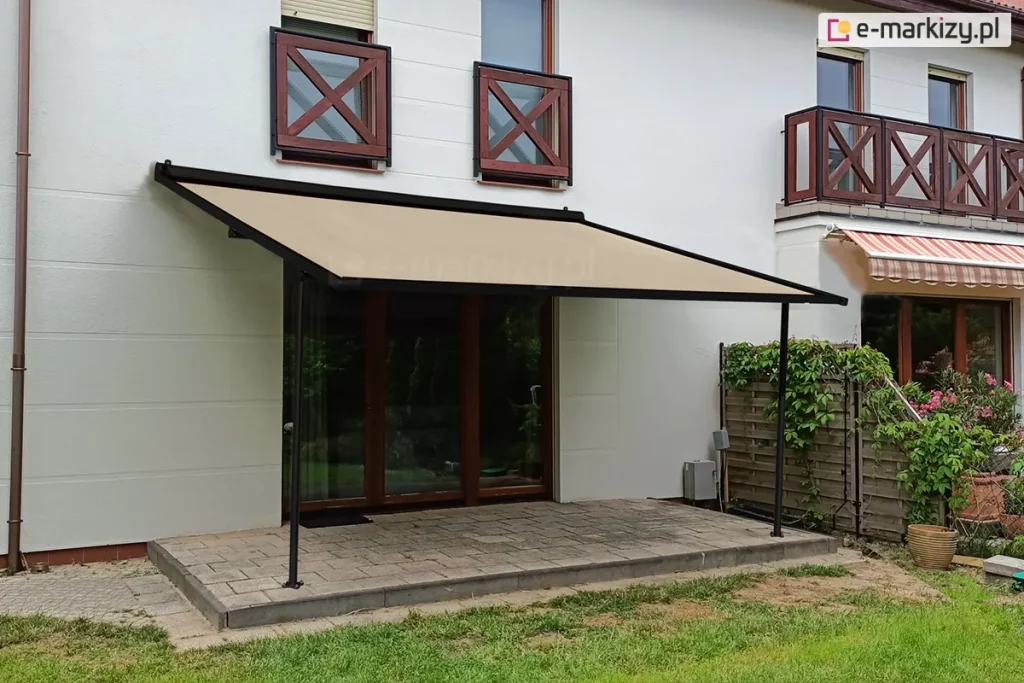 Nowoczesna pergola tarasowa przyścienna Vera-Sola o lekkiej konstrukcji aluminiowej w kolorze czarnym, zamontowana do białej elewacji piętrowego domu jednorodzinnego. Zadaszenie stanowi gładkie poszycie tkaninowe w kolorze beżowym, które jest całkowicie rozłożone pod kątem, wspierając się na dwóch smukłych profilach pionowych zamocowanych do podłoża. Pergola osłania taras wyłożony szarymi, prostokątnymi płytami chodnikowymi, na który prowadzą przeszklone drzwi tarasowe. Po prawej stronie konstrukcji znajduje się drewniane ogrodzenie panelowe częściowo porośnięte zielonymi pnączami oraz donice z roślinnością, a w tle widoczny jest fragment sąsiedniego balkonu z brązową barierką i wysuniętą markizą w czerwono-beżowe pasy. Całość znajduje się na tle zadbanego trawnika w pełnym świetle dziennym, prezentując minimalistyczne podejście do ochrony przeciwsłonecznej strefy wypoczynkowej.
