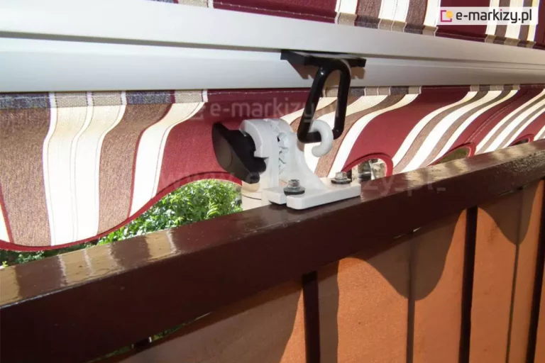 Barierka balkonu z w kolorze brązowym z przesłoną tkaninową o brązowo-pomarańczowym odcieniu zamocowaną w balustradzie. Do powierzchni barierki przymocowane białe ramię markizy poręczowej z widocznymi dwoma ocynkowanymi śrubami oraz czarnym pokrętłem do zmiany kąta nachylenia ramienia. Ponad śrubami widoczny biały zawijas, o który zahaczono czarny zaczep oczkowy, przymocowany rowku białej belki przedniej markizy. Poniżej belki przedniej widoczna falowana falbana w bordowe, brązowe i kremowo-białe pasy, spod której widoczne są zielone krzewy.