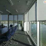 Wnętrze nowoczesnej sali konferencyjnej z przeszkleniami na całą wysokość ścian, wyposażonymi w system wewnętrznych rolet przeciwsłonecznych typu screen. Refleksol XL Aluprof o jasnoszarym odcieniu i wysokim stopniu przezierności są częściowo opuszczone, co pozwala na redukcję odblasków przy jednoczesnym zachowaniu widoku na jezioro i zieleń za oknem. W pomieszczeniu znajduje się długi, ciemny stół konferencyjny z niebieskimi fotelami biurowymi na kółkach oraz rośliny doniczkowe ustawione przy oknach. Na suficie widoczne jest profesjonalne oświetlenie liniowe, punktowe oraz rzutnik multimedialny. Przeszklone ściany odbijają naturalne światło dzienne, a na podłodze z ciemną wykładziną widoczne są kratki wentylacyjne i puszki podłogowe.
