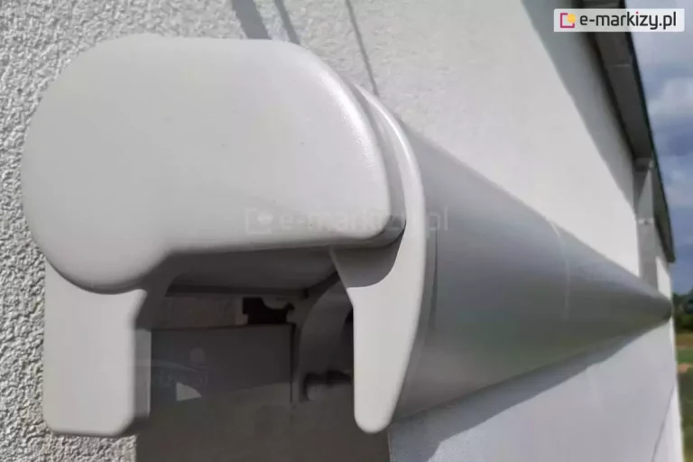 Markiza Palladio Aluprof ukazana od boku kasety w srebrnym kolorze. Wyraźnie widoczna zaślepka boczna z pcv oraz belka przednia markizy z zamocowaną w niej falbaną. Markiza zawieszona na ścianie z jasnym tynkiem elewacji.