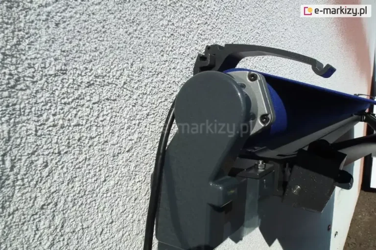Markiza bez kasety w kolorze grafitowym zawieszona na ścianie z białym tynkiem strukturalnym z widocznym kablem zasilania napędu elektrycznego oraz wspornikiem do uniwersalnego daszka do markiz, na którym opiera się górna blacha osłony
