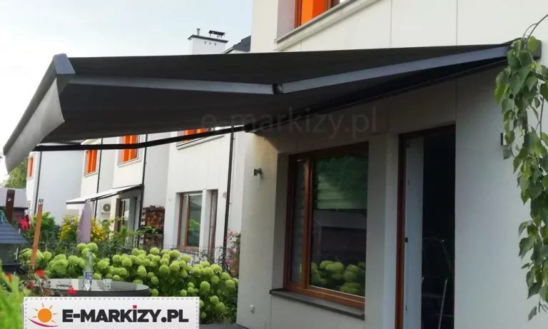 Zdjęcie prezentuje nowoczesną markizę tarasową Moderno MOL w pełnej kasecie, zamontowaną na jasnej elewacji nowoczesnego budynku szeregowego. Na obrazku Markiza Moderno MOL Galeria Zdjęć markiza jest całkowicie rozłożona, a jej poszycie oraz konstrukcja (ramiona przegubowe i listwa frontowa) wykonane są w eleganckim, ciemnym kolorze antracytowym. Materiał osłony jest gładki i precyzyjnie napięty, tworząc szeroki dach nad tarasem na parterze. W tle widoczne są sąsiednie segmenty białych budynków z charakterystycznymi pomarańczowymi akcentami w oknach. Bezpośrednio pod markizą znajduje się okno w drewnianej ramie oraz fragment ogrodu z gęstymi, zielonymi krzewami hortensji i okrągłym stolikiem kawowym. Całość ujęcia, wykonana przy naturalnym świetle dziennym, podkreśla minimalistyczny design produktu oraz jego funkcjonalność w zabudowie jednorodzinnej.