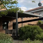 Aluminiowa pergola tarasowa Square w kolorze ciemnego antracytu, zamontowana do ściany budynku wykonanej z czerwonej cegły klinkierowej. Konstrukcja zadaszenia opiera się na poprzecznych profilach, na których rozpięta jest jasna, kremowa tkanina układająca się w regularne pasy. W profilach zadaszenia zamontowano punktowe oświetlenie LED, które rzuca delikatne światło na przestrzeń poniżej. Całość wspiera się na solidnym, pionowym słupie o kwadratowym przekroju, a z boku widoczna jest pozioma belka wzmacniająca konstrukcję. Pod pergolą, na podwyższonym tarasie, znajduje się zestaw mebli ogrodowych, w tym krzesła z szarej plecionki ustawione przy stole. Taras jest otoczony gęstą, naturalną roślinnością – na pierwszym planie oraz po prawej stronie widoczne są rozłożyste, zielone krzewy, które częściowo przesłaniają widok na meble. W górnej części zdjęcia zwieszają się gałęzie drzewa liściastego z wyraźnie zarysowanymi zielonymi liśćmi. W tle widać fragment sąsiedniego piętrowego domu o jasnej elewacji z ciemnoczerwonym dachem, na którym zamontowana jest antena satelitarna. Niebo jest zachmurzone, w odcieniach szarości i błękitu, co nadaje zdjęciu naturalny, dzienny charakter.