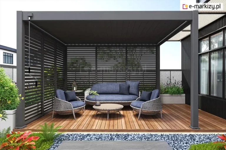 Nowoczesna pergola tarasowa w kolorze antracytowym, zainstalowana na drewnianym podeście z brązowych desek kompozytowych. Konstrukcja posiada dach lamelowy oraz system ścianek bocznych w formie nieruchomych żaluzji zewnętrznych, które osłaniają dwa boki altany, zapewniając ochronę przed słońcem i wiatrem. Przy słupie z lewej strony widoczna korba do ręcznego sterowania kątem pochylenia lamel dachowych. Wewnątrz strefy wypoczynkowej znajduje się zestaw mebli ogrodowych o ozdobnym splocie, składający się z trzyosobowej sofy, dwóch foteli z granatowymi poduszkami oraz niskiego, okrągłego stolika kawowego. Przed tarasem znajduje się pas ozdobnego, ciemnego żwiru oraz niska roślinność ogrodowa, w tym paprocie i kwiaty o barwnych liściach. W tle widoczna jest elewacja nowoczesnego budynku z dużymi przeszkleniami oraz fragment sąsiedniej zabudowy, co podkreśla miejski charakter aranżacji.