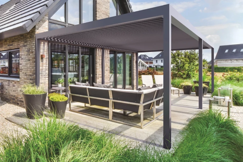 Nowoczesna pergola lamelowa wolnostojąca SB450 marki Aluprof w kolorze ciemnoszarym, zamontowana jako zadaszenie tarasu przy piętrowym domu jednorodzinnym. Konstrukcja pergoli opiera się na pionowych, masywnych słupach o kwadratowym przekroju i prostej ramie górnej, w której osadzono dach z ruchomych, aluminiowych lameli. Elewacja budynku wykonana jest z klinkierowej cegły w odcieniach brązu i beżu, z dużymi, przeszklonymi drzwiami tarasowymi w ciemnych ramach, w których odbija się otoczenie. Pod pergolą, na jasnej, drewnianej posadzce tarasowej, ustawiono luksusowy komplet wypoczynkowy składający się z dużej, narożnej kanapy z ciemnoszarymi poduszkami na metalowym stelażu oraz leżaków ogrodowych w tle. Wokół tarasu zaaranżowano nowoczesny ogród z gęstymi kępami ozdobnych, zielonych traw posadzonych w gruncie oraz w dużych, ciemnoszarych donicach stożkowych. W tle widoczne są inne domy jednorodzinne, drzewa oraz jasne, lekko zachmurzone niebo w świetle dziennym.