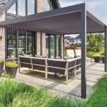 Nowoczesna pergola lamelowa wolnostojąca SB450 marki Aluprof w kolorze ciemnoszarym, zamontowana jako zadaszenie tarasu przy piętrowym domu jednorodzinnym. Konstrukcja pergoli opiera się na pionowych, masywnych słupach o kwadratowym przekroju i prostej ramie górnej, w której osadzono dach z ruchomych, aluminiowych lameli. Elewacja budynku wykonana jest z klinkierowej cegły w odcieniach brązu i beżu, z dużymi, przeszklonymi drzwiami tarasowymi w ciemnych ramach, w których odbija się otoczenie. Pod pergolą, na jasnej, drewnianej posadzce tarasowej, ustawiono luksusowy komplet wypoczynkowy składający się z dużej, narożnej kanapy z ciemnoszarymi poduszkami na metalowym stelażu oraz leżaków ogrodowych w tle. Wokół tarasu zaaranżowano nowoczesny ogród z gęstymi kępami ozdobnych, zielonych traw posadzonych w gruncie oraz w dużych, ciemnoszarych donicach stożkowych. W tle widoczne są inne domy jednorodzinne, drzewa oraz jasne, lekko zachmurzone niebo w świetle dziennym.