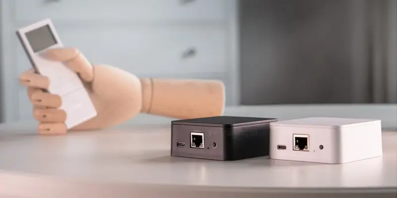 Zbliżenie na zestaw urządzeń systemu inteligentnego domu Yoda Smart Home 3, w skład którego wchodzą dwie centrale sterujące w formie niewielkich, prostopadłościennych skrzynek o zaokrąglonych krawędziach, ustawionych obok siebie na blacie stołu. Jedna z jednostek centralnych posiada obudowę w kolorze czarnym, natomiast druga w kolorze białym, przy czym obie zostały wyposażone w identyczny panel złączy umieszczony na przedniej ściance, obejmujący port USB typu C, gniazdo Ethernet RJ-45, diodę sygnalizacyjną oraz fizyczny przycisk resetu. W tle, nieco nieostro, widoczna jest drewniana dłoń modelarska wykonana z jasnego drewna, która w zgiętych palcach trzyma biały, prostokątny pilot zdalnego sterowania z wyświetlaczem, co sugeruje proces konfiguracji lub obsługi automatyki domowej. Całość kompozycji utrzymana jest w jasnej, stonowanej kolorystyce, skupiając uwagę na urządzeniach sieciowych i ergonomicznym akcesorium sterującym, podkreślając nowoczesny charakter technologii smart home oraz jej łatwość w obsłudze.