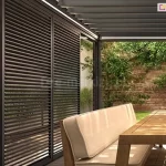 Wizualizacja przedstawia nowoczesną pergolę tarasową z przesuwnymi ścianami bocznymi w formie aluminiowych okiennic żaluzjowych w kolorze ciemnego antracytu lub czarnym. Dwa wysokie, przesuwne panele z poziomymi, stałymi lamelami tworzą ażurową przesłonę, zapewniającą ochronę przed słońcem, wiatrem oraz prywatność. Panele te poruszają się po górnej prowadnicy zamontowanej w belce konstrukcyjnej pergoli. Dach pergoli składa się z obrotowych lameli w tym samym ciemnym kolorze, a pod belkami obwodowymi zintegrowane jest oświetlenie liniowe LED, które oświetla przestrzeń pod zadaszeniem. Wewnątrz pergoli, na podłodze wyłożonej jasnym gresem, stoi drewniany stół jadalniany z jasnym blatem, a przy nim sofa tarasowa z beżowymi poduszkami siedziskowymi i oparciowymi. Na stole umieszczono dwie ozdobne, ryflowane wazony w kształcie kuli w odcieniach złota/brązu. W tle, za pergolą, widoczna jest ściana z surowej, czerwonej cegły z rosnącymi na niej pnączami i krzewami. Po lewej stronie, na jasnej ścianie elewacyjnej, zamontowany jest nowoczesny, czarny kinkiet zewnętrzny o minimalistycznym designie. Wzdłuż ściany ciągnie się pas ozdobnych traw i roślin zielonych w betonowych donicach, a podłoga przechodzi w podest z desek kompozytowych. Całość kompozycji łączy industrialny charakter metalowej konstrukcji z ciepłem drewna i naturalną zielenią ogrodu, tworząc funkcjonalną i estetyczną strefę wypoczynkową.