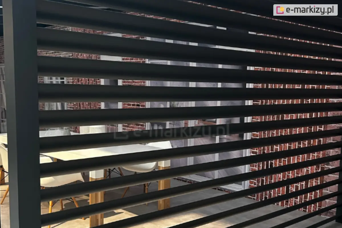 Zbliżenie na boczny panel żaluzjowy Louvre zamontowany w konstrukcji nowoczesnej pergoli tarasowej. Zdjęcie prezentuje detale techniczne poziomych lameli wykonanych z aluminium w kolorze ciemnego grafitu, które są rozmieszczone w regularnych odstępach, tworząc ażurową przesłonę. Przez szczeliny panelu żaluzjowego Louvre widoczne jest tło przedstawiające fragment tarasu z meblami jadalnianymi – białym stołem na drewnianych nogach oraz jasnymi krzesłami. Za meblami znajduje się ściana budynku wykonana z czerwonej cegły z dużymi przeszkleniami. Lewa krawędź zdjęcia ukazuje pionowy profil nośny pergoli, do którego przymocowany jest panel żaluzjowy Louvre. Światło dzienne wpadające między lamele tworzy rytmiczny wzór linii i cieni, podkreślając matowe wykończenie i precyzję wykonania elementów żaluzji.