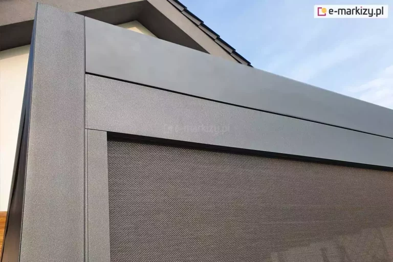 zbliżenie na detale konstrukcyjne narożnika pergoli tarasowej Luxury wykonanej z aluminium. Konstrukcja jest wykończona lakierem proszkowym o drobnej strukturze w kolorze ciemnego antracytu. W górnej części widoczny jest szeroki, płaski profil czołowy, pod którym zamontowano kasetę refleksola Zip chroniącą mechanizm zwijający tkaniny rolety bocznej. Poniżej profilu znajduje się zintegrowana prowadnica pionowa, w której umieszczona jest tkanina rolety zewnętrznej typu screen. Materiał rolety ma gęsty splot w kolorze grafitowym lub ciemnobrązowym i jest precyzyjnie napięty wewnątrz ramy. W tle widać fragment jasnej ściany budynku oraz krawędź dachu z ciemną dachówką pod jasnym niebem. Fotografia ukazuje wysoką jakość spasowania elementów aluminiowych oraz estetykę nowoczesnej zabudowy tarasu na wymiar.