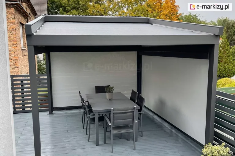 Wolnostojąca pergola tarasowa Suare o konstrukcji aluminiowej w kolorze ciemnego antracytu, posadowiona na podwyższonym tarasie wykonanym z gładkich desek kompozytowych w jasnoszarym kolorze. Zadaszenie pergoli stanowi jednolity, napięty materiał w kolorze szarym, który jest lekko nachylony w jedną stronę, aby zapewnić odpływ wody. Dwie ściany konstrukcji – tylna oraz boczna po prawej stronie – zostały całkowicie przesłonięte pionowymi osłonami typu screen w jasnoszarym odcieniu, co tworzy intymną i osłoniętą od wiatru przestrzeń. Wewnątrz pergoli znajduje się strefa jadalniana z dużym, prostokątnym stołem w kolorze antracytowym oraz sześcioma dopasowanymi krzesłami z tekstylnym obiciem. Na środku stołu ustawiono małą doniczkę z rośliną ozdobną. Taras ograniczony jest nowoczesną balustradą z poziomych, szerokich profili metalowych w kolorze ciemnoszarym. W bezpośrednim sąsiedztwie widoczny jest fragment ściany starego budynku z czerwonej cegły oraz wysokie drzewa liściaste o jesiennym, żółto-pomarańczowym zabarwieniu liści. Po prawej stronie w tle rysuje się linia zielonych drzew iglastych pod jasnym, lekko zachmurzonym niebem.