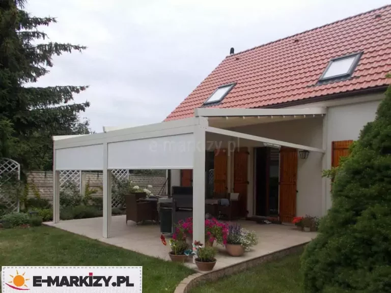 Nowoczesna pergola przyścienna Moderno o konstrukcji w kolorze kremowym, zamontowana przy budynku z jasną elewacją i dachem z czerwonej dachówki. Zadaszenie tkaninowe jest w pełni rozwinięte, tworząc osłonę nad jasnym tarasem, na którym ustawiono zestaw mebli obiadowych z ciemnego technorattanu. Frontowa część pergoli została wyposażona w dwie pionowe przesłony typu refleksol w pasującym jasnym kolorze, które są częściowo opuszczone, zapewniając ochronę przed słońcem. Aranżację uzupełniają liczne kwiaty w donicach ustawione na krawędzi tarasu oraz widoczna w tle zieleń ogrodowa z drewnianym ogrodzeniem i pergolami kratkowymi.