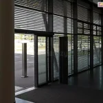 Widok z wnętrza nowoczesnego holu budynku na przeszkloną ścianę frontową z zainstalowanymi żaluzjami fasadowymi Aluprof. Poziome lamele żaluzji są ustawione w pozycji półotwartej, co pozwala na przenikanie światła dziennego do środka, tworząc rytmiczny wzór cieni na podłodze, jednocześnie ograniczając nagrzewanie pomieszczenia. Przez przeszklenia widoczny jest plac zewnętrzny oraz fragmenty zieleni miejskiej. Konstrukcja żaluzji obejmuje całą wysokość witryn, od podłogi aż po sufit, z wyraźnie zaznaczonymi podziałami pionowymi ram okiennych i drzwi wejściowych. Po lewej stronie kadru widoczny jest masywny filar konstrukcyjny budynku, a wewnątrz znajdują się elementy infrastruktury, takie jak ciemna wycieraczka systemowa i metalowe słupki informacyjne.