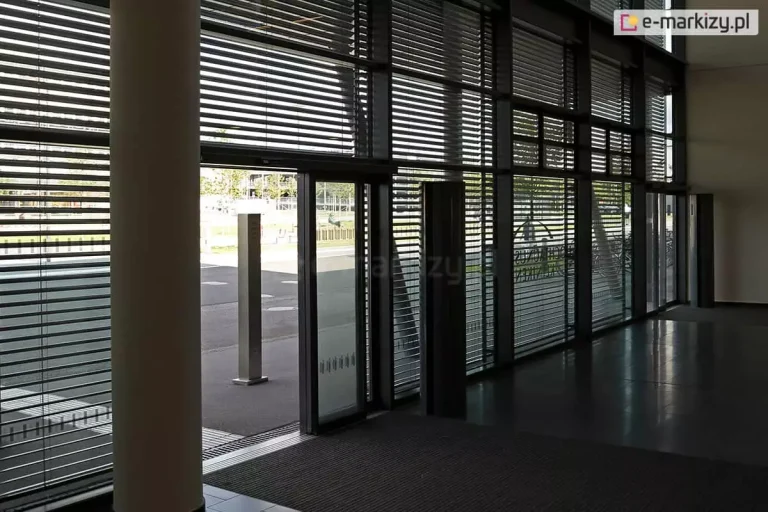 Widok z wnętrza nowoczesnego holu budynku na przeszkloną ścianę frontową z zainstalowanymi żaluzjami fasadowymi Aluprof. Poziome lamele żaluzji są ustawione w pozycji półotwartej, co pozwala na przenikanie światła dziennego do środka, tworząc rytmiczny wzór cieni na podłodze, jednocześnie ograniczając nagrzewanie pomieszczenia. Przez przeszklenia widoczny jest plac zewnętrzny oraz fragmenty zieleni miejskiej. Konstrukcja żaluzji obejmuje całą wysokość witryn, od podłogi aż po sufit, z wyraźnie zaznaczonymi podziałami pionowymi ram okiennych i drzwi wejściowych. Po lewej stronie kadru widoczny jest masywny filar konstrukcyjny budynku, a wewnątrz znajdują się elementy infrastruktury, takie jak ciemna wycieraczka systemowa i metalowe słupki informacyjne.