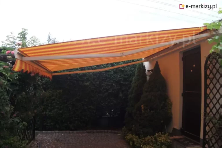 Zdjęcie prezentuje w pełni rozłożoną markizę tarasową Palladio Aluprof, która osłania przydomowe miejsce wypoczynku wyłożone kostką brukową. Poszycie markizy wykonane jest z tkaniny w intensywne, pionowe pasy w odcieniach żółci, pomarańczu oraz czerwieni, co nadaje instalacji ciepły charakter. Przednia krawędź materiału zakończona jest dekoracyjną, falowaną falbaną, która powiela pasiasty wzór poszycia. Konstrukcja wsporcza, obejmująca ramiona przegubowe oraz belkę frontową, jest w kolorze białym, co kontrastuje z barwnym materiałem. Markiza jest zamontowana do bocznej ściany budynku o jasnym, kremowym tynku. Tuż pod osłoną znajdują się ciemnobrązowe drzwi wejściowe oraz gęsta roślinność ogrodowa, w tym wysokie, przycięte w stożek iglaki. Po lewej stronie kadru widoczny jest fragment pergoli lub trejażu, po którym pną się krzewy róż z widocznymi kwiatami w kolorze różowym. Tło stanowi wysoki, gęsty żywopłot, który szczelnie odgradza posesję, zapewniając prywatność. Oświetlenie na zdjęciu sugeruje pochmurny dzień lub miejsce znajdujące się w głębokim cieniu, co podkreśla funkcję ochronną markizy przed warunkami atmosferycznymi.