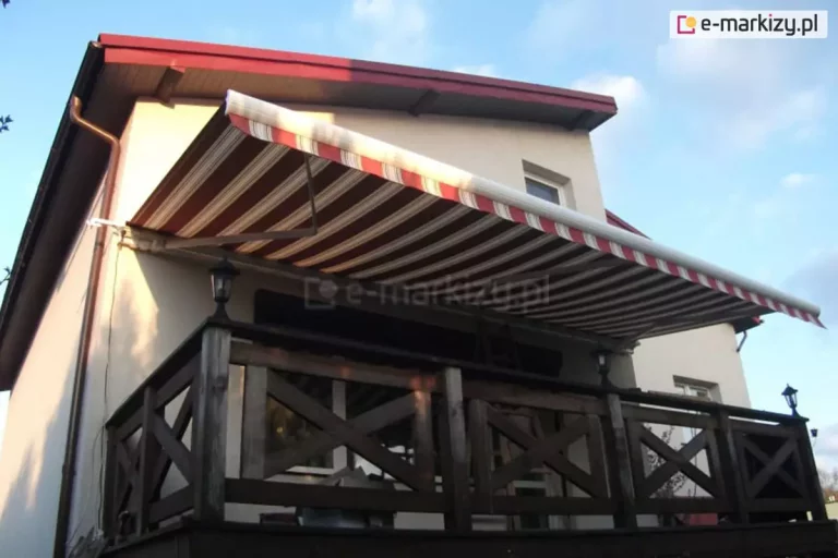 Zdjęcie prezentuje szeroką markizę tarasową Palladio Aluprof zamontowaną na elewacji piętrowego domu jednorodzinnego, osłaniającą przestronny balkon. Poszycie markizy wykonane jest z materiału akrylowego w klasyczne, poziome pasy w kolorystyce bordowej, brązowej i beżowej, co harmonizuje z ciemną stolarką oraz dachem budynku. Markiza jest w pełni rozwinięta, wsparta na białych ramionach przegubowych zamontowanych do jasnej, tynkowanej ściany. Balkon zabezpieczony jest solidną, drewnianą barierką o ciemnobrązowym odcieniu, charakteryzującą się konstrukcją z krzyżującymi się belkami wypełniającymi poszczególne przęsła. Na balustradzie zamontowane są klasyczne, czarne lampiony zewnętrzne pełniące funkcję oświetlenia. Budynek posiada dwuspadowy dach o ciemnoczerwonym pokryciu z widocznymi podbitkami dachowymi w kolorze naturalnego drewna. Po lewej stronie fasady widoczna jest miedziana rura spustowa odprowadzająca wodę z rynny. Całość kompozycji uchwycona jest z dolnej perspektywy na tle błękitnego nieba z delikatnymi chmurami, co podkreśla funkcjonalność osłony przeciwsłonecznej w słoneczny dzień.