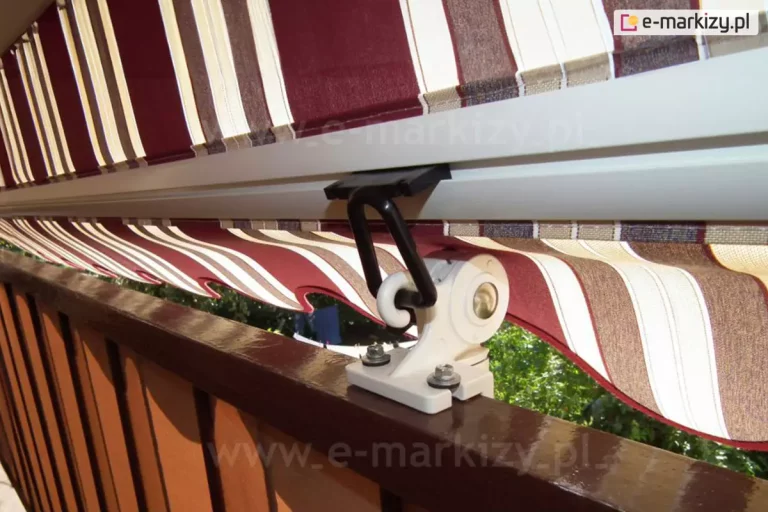Markiza balkonowa z ramionami poręczowymi zamocowana do haczyka uchwytu ramienia mocowanego do barierki balkonu za pomocą zaczepu oczkowego wykonanego z czarnego PCV. Zaczep oczkowy do ramion Italia poręczowa wsunięty jest w specjalnie wykonany rowek białej belki przedniej markizy. Całość zamocowana na balkonie o brązowej barierce, markizę wyposażono w poszycie oraz falowaną falbanę wykonaną z tkaniny akrylowej w bordowe, kremowe i białe pasy.