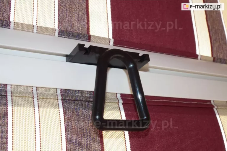 Czarny zaczep oczkowy do ramion poręczowych wykonany z tworzywa PCV wsunięty w rowek aluminiowej belki przedniej markizy balkonowej z ramionami poręczowymi w kolorze białym, widoczny na tle tkaniny poszycia w bordowe, kremowe i białe pasy.