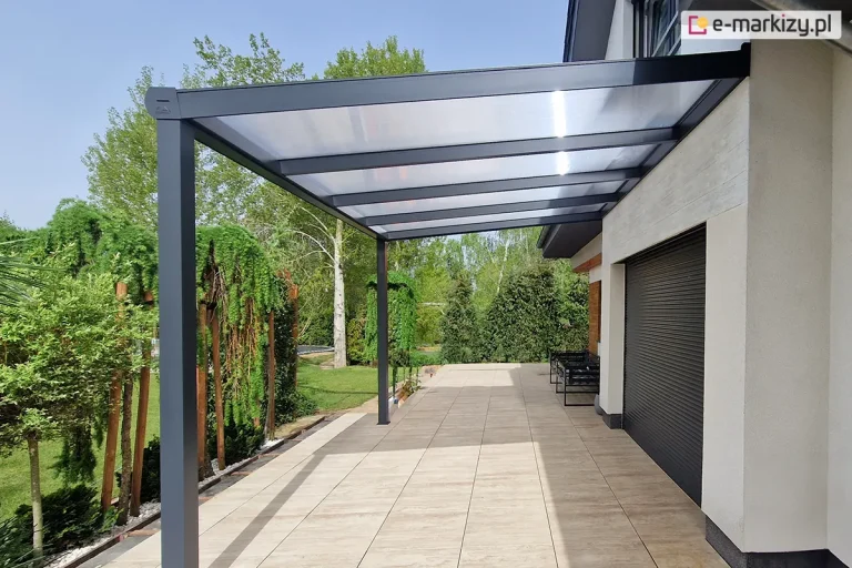 Boczne ujęcie nowoczesnego zadaszenia tarasowego Gumax (obecnie Deponti) o konstrukcji w kolorze antracytowym, które zostało zamontowane do jasnej elewacji piętrowego domu. Aluminiowa pergola posiada jednospadowy dach z wypełnieniem z mlecznych, półprzezroczystych paneli poliwęglanowych, wsparty na dwóch solidnych, kwadratowych słupach nośnych. Pod zadaszeniem znajduje się przestronny taras wyłożony beżowymi płytami o teksturze drewna, na którym w głębi ustawiono czarną, metalową ławkę. Na ścianie budynku widoczna jest duża, ciemnoszara roleta zewnętrzna oraz ozdobny fragment elewacji z drewnopodobnych paneli. Otoczenie stanowi gęsty ogród z wysokimi brzozami, krzewami oraz fragmentem iglaka na pierwszym planie, a całość dopełnia bezchmurne, jasne niebo.