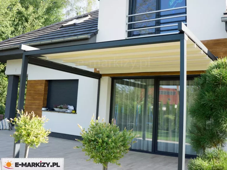 Zdjęcie prezentuje nowoczesne zadaszenie tarasowe Luxo o konstrukcji aluminiowej w kolorze antracytowym, zamontowane do białej elewacji piętrowego budynku. Pergola posiada w pełni rozwinięty dach wykonany z jasnej, kremowej tkaniny technicznej, która jest rozpięta na poprzecznych profilach nośnych. Konstrukcja wspiera się na pionowych słupach o kwadratowym przekroju, osadzonych na tarasie wyłożonym jasnoszarymi płytami gresowymi. Na elewacji domu widoczne są ozdobne panele z naturalnego drewna oraz duże przeszklenia z ciemnymi ramami, w których odbija się ogród. Na tarasie, obok słupów pergoli, ustawiono rośliny ozdobne w formie niewielkich drzewek o kulistych koronach oraz krzewy iglaste. W tle, za dużym oknem tarasowym, dostrzec można jasne zasłony, a powyżej fragment balkonu z metalową barierką oraz spadzisty dach pokryty ciemną dachówką z oknem połaciowym. Całość ukazuje minimalistyczną i elegancką aranżację zewnętrznej strefy relaksu, zintegrowaną z nowoczesną architekturą domu.