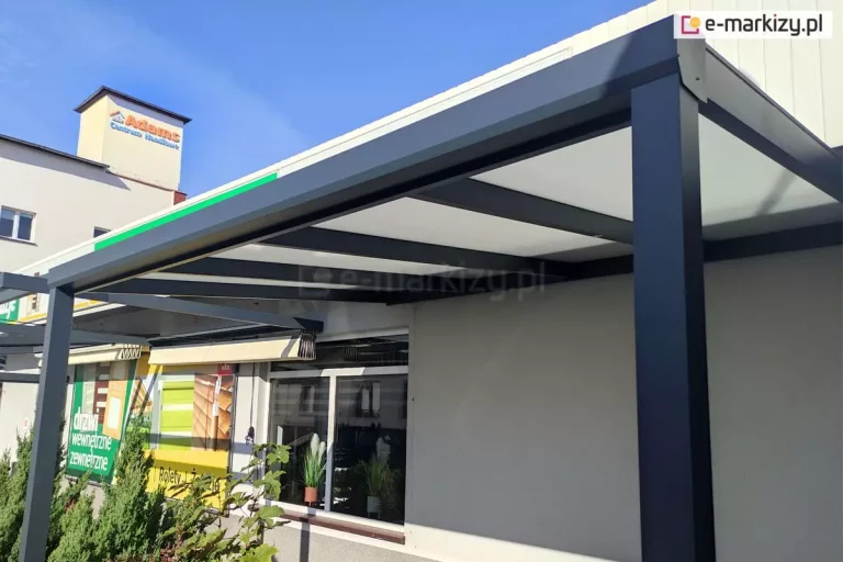Pergola aluminiowa przyścienna z nowoczesnym zadaszeniem z poliwęglanu komorowego zamontowana przy budynku. Solidna konstrukcja w kolorze antracytowym tworzy eleganckie zadaszenie tarasu przed lokalem handlowym w słoneczny dzień. W tle widoczne duże okno salonu sprzedaży oraz baner reklamowy i dwie inne pergole. Nad wszystkim widoczny komin sąsiedniego budynku z logo sklepu "Adams centrum handlowe" na tle błękitnego nieba.