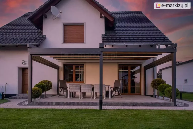 Nowoczesną, przyścienna pergola tarasowa Solid zamontowana przy domu jednorodzinnym, uchwyconą o zmierzchu przy efektownym, purpurowym niebie. Aluminiowa konstrukcja dwóch modułów, w kolorze ciemnego antracytu posiada dach tkaninowy, który jest częściowo rozsunięty, a pod jego poszyciem widoczne są zintegrowane, liniowe paski LED emitujące ciepłe, żółte światło. Pod zadaszeniem, na tarasie wyłożonym szarymi płytami, znajduje się strefa jadalniana z dużym stołem i jasnymi, plecionymi krzesłami. Budynek ma jasną elewację, ciemną dachówkę i okna w drewnianych ramach. Całość otacza zadbany trawnik oraz symetrycznie posadzone, formowane w kule krzewy, co nadaje przestrzeni elegancki i przytulny charakter.