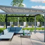 Zdjęcie prezentuje produkt pergola tarasowa wolnostojąca Moderno usytuowaną w nowoczesnej aranżacji ogrodowej przy basenie. Konstrukcja pergoli charakteryzuje się minimalistyczną formą, wykonaną z solidnych profili aluminiowych o kwadratowym przekroju w kolorze głębokiego antracytu. Zadaszenie stanowi jasny, kremowy materiał techniczny rozpięty na ruchomych poprzeczkach, który jest zamontowany pod lekkim skosem, co ułatwia odprowadzanie wody opadowej. Pod pergolą znajduje się strefa wypoczynkowa z dużym, narożnym meblem modułowym w kolorze jasnobłękitnym, ustawionym na podłożu z jasnych, wielkoformatowych płyt tarasowych. Obok sofy stoi niski, nowoczesny stolik kawowy z szklanym blatem, na którym umieszczono dekoracyjne naczynia. Tło kompozycji stanowi zadbany ogród z zielonym trawnikiem, niską roślinnością ozdobną oraz nowoczesnym ogrodzeniem panelowym w kolorze czarnym z betonowymi słupkami. Po prawej stronie kadru widoczna jest krawędź błękitnej tafli wody basenowej, a w oddali gęsta ściana drzew liściastych pod jasnym, lekko zachmurzonym niebem. Cała scena jest oświetlona naturalnym, dziennym światłem, podkreślającym detale wykończenia pergoli oraz fakturę materiałów.