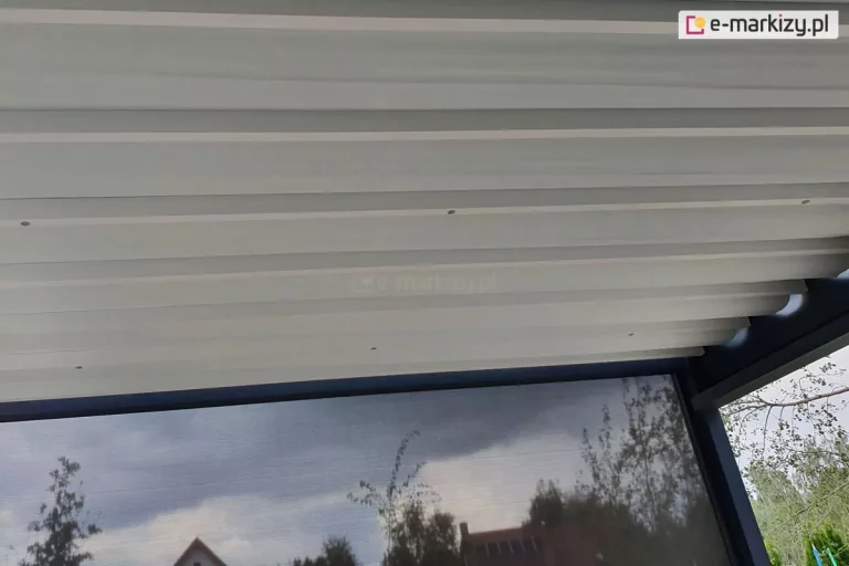 Zdjęcie prezentuje widok od dołu na rozłożone zadaszenie tkaninowe nowoczesnej pergoli tarasowej Luxury z tkaniny Opatex w kolorze jasnokremowym. Materiał zadaszenia jest rozpięty na poprzecznych profilach aluminiowych, tworząc regularne, poziome segmenty. Poniżej linii dachu, po prawej stronie, widoczna jest opuszczona roleta boczna typu screen w kolorze ciemnoszarym, która jest półprzezroczysta i odbija zarysy drzew oraz pobliskich budynków pod zachmurzonym niebem. Konstrukcja nośna pergoli wykonana jest z aluminium lakierowanego proszkowo na matowy kolor antracytowy. W prawym dolnym rogu kadru widać fragmenty zielonych koron drzew, co sugeruje umiejscowienie tarasu w ogrodzie. Perspektywa zdjęcia podkreśla estetyczne połączenie jasnej tkaniny dachowej z ciemną ramą konstrukcji oraz funkcjonalność zintegrowanego systemu osłon przeciwsłonecznych.