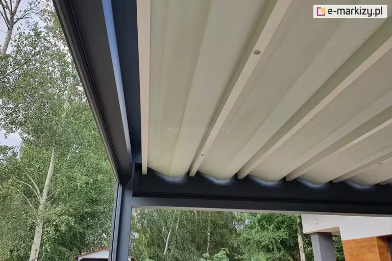 Zdjęcie przedstawia widok od dołu na rozłożone zadaszenie tkaninowe nowoczesnej pergoli tarasowej Luxury. Poszycie wykonane jest z jasnej, kremowej tkaniny wodoodpornej, która jest rozpięta na poprzecznych profilach aluminiowych. Materiał ułożony jest w charakterystyczne, lekko falujące segmenty. Główna rama pergoli jest wykonana w kolorze ciemnego antracytu, co tworzy wyraźny kontrast z jasnym poszyciem i białymi belkami przeciwwietrznymi. Po lewej stronie kadru widoczny jest pionowy profil konstrukcyjny oraz fragment ogrodu z wysokimi, zielonymi drzewami liściastymi na tle zachmurzonego nieba. W prawym dolnym rogu widać fragment jasnej elewacji budynku z elementami wykończonymi drewnem. Perspektywa zdjęcia podkreśla techniczne aspekty odprowadzania wody oraz estetykę wykończenia spodu zadaszenia.