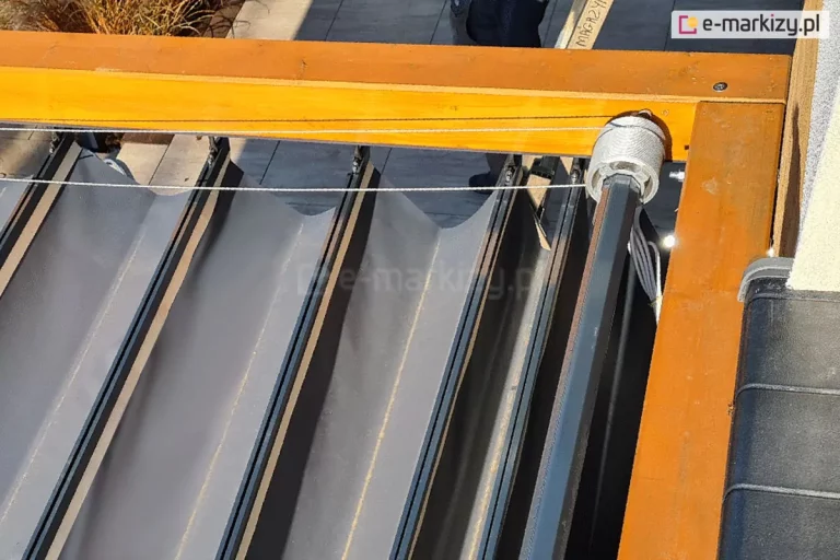 Zadaszenie rzymskie supro z napędem elektrycznym zamontowane nad tarasem na pergoli drewnianej Juko. Widoczny system sterowania oraz tkanina Opatex w kolorze antracytowym zamocowana do belek przeciwwietrznych.