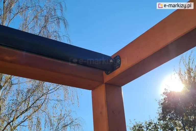 Narożnik konstrukcji drewnianej pergoli tarasowej z zamocowaną do jednej z belek prowadnicą dachu Silva w kolorze antracytowym. W tle widoczne drzewa oraz błękitne niebo i przeświecające zza liści słońce.