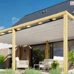 Zadaszenie tarasu drewniane juko wolnostojąca pergola ustawiona przy ścianie domu piętrowego o drewnianej fasadzie. W tle widoczny żywopłot i niebieskie niebo z malutkimi białymi chmureczkami.