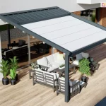 Nowoczesna pergola przyścienna Moderno o konstrukcji aluminiowej w kolorze antracytowym, zamontowana nad przestronnym tarasem wyłożonym deskami o naturalnym odcieniu drewna. Zadaszenie składa się z dwóch części: stałego daszka systemowego w kolorze antracytowym chroniącego zwinięty materiał oraz rozwiniętej, jasnej tkaniny technicznej podzielonej na równe segmenty. Pod pergolą zaaranżowano strefę wypoczynkową z szarymi meblami tarasowymi o konstrukcji drewnianej, białymi poduszkami, stolikami kawowymi oraz roślinami w ozdobnych czarnych donicach. Konstrukcja przylega do białej elewacji budynku z dużym przeszkleniem, w którym odbija się otoczenie, a w tle widoczny jest fragment ściany z czerwonej cegły i dekoracyjna misa z roślinnością.