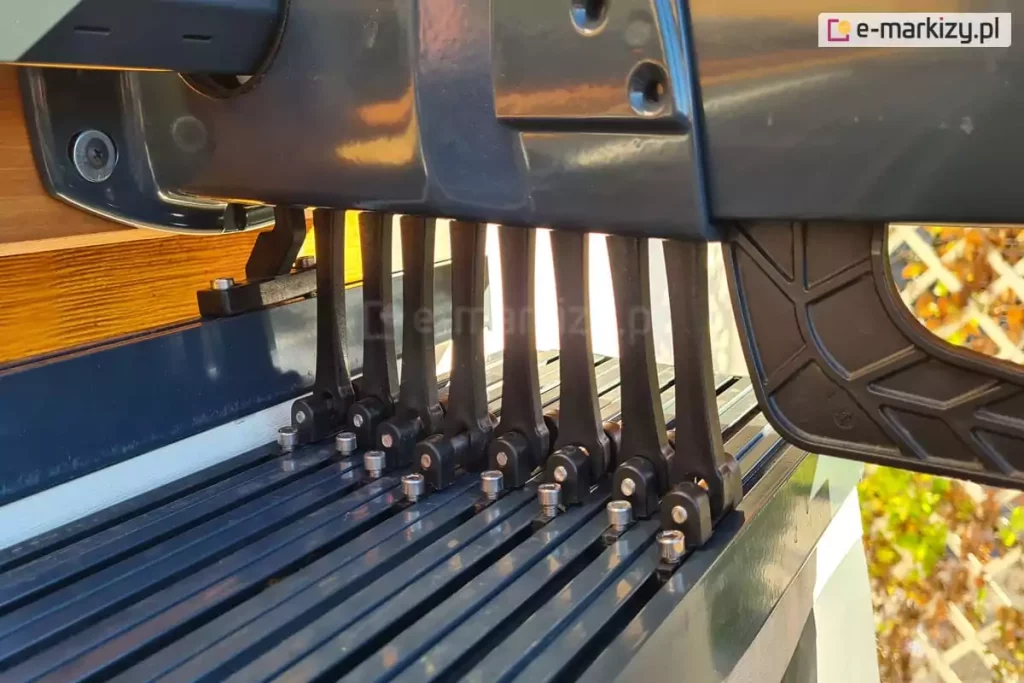Zdjęcie prezentuje zbliżenie na mechanizm prowadzący ruchomego zadaszenia rzymskiego do pergoli model Silva w kolorze antracytowym. Widoczny jest szereg czarnych, pionowych wózków jezdnych zamontowanych na szynach jezdnych, które odpowiadają za podtrzymywanie i przesuwanie profili tkaniny. Każdy wózek jest przymocowany do listwy przeciwwietrznej za pomocą srebrnych śrub imbusowych. Nad mechanizmem znajduje się masywna, aluminiowa obudowa prowadnicy o gładkiej, błyszczącej powierzchni. Po lewej stronie widoczny jest fragment drewnianej belki konstrukcyjnej o wyraźnym usłojeniu oraz element mocujący prowadnicę do stelaża. W tle, poza ostrością, widoczna jest jasna elewacja budynku oraz ażurowa kratka ogrodowa z roślinnością. Zdjęcie szczegółowo ukazuje techniczną stronę systemu przesuwnego, podkreślając precyzję wykonania elementów mechanicznych.