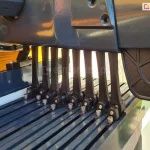 Zdjęcie prezentuje zbliżenie na mechanizm prowadzący ruchomego zadaszenia rzymskiego do pergoli model Silva w kolorze antracytowym. Widoczny jest szereg czarnych, pionowych wózków jezdnych zamontowanych na szynach jezdnych, które odpowiadają za podtrzymywanie i przesuwanie profili tkaniny. Każdy wózek jest przymocowany do listwy przeciwwietrznej za pomocą srebrnych śrub imbusowych. Nad mechanizmem znajduje się masywna, aluminiowa obudowa prowadnicy o gładkiej, błyszczącej powierzchni. Po lewej stronie widoczny jest fragment drewnianej belki konstrukcyjnej o wyraźnym usłojeniu oraz element mocujący prowadnicę do stelaża. W tle, poza ostrością, widoczna jest jasna elewacja budynku oraz ażurowa kratka ogrodowa z roślinnością. Zdjęcie szczegółowo ukazuje techniczną stronę systemu przesuwnego, podkreślając precyzję wykonania elementów mechanicznych.