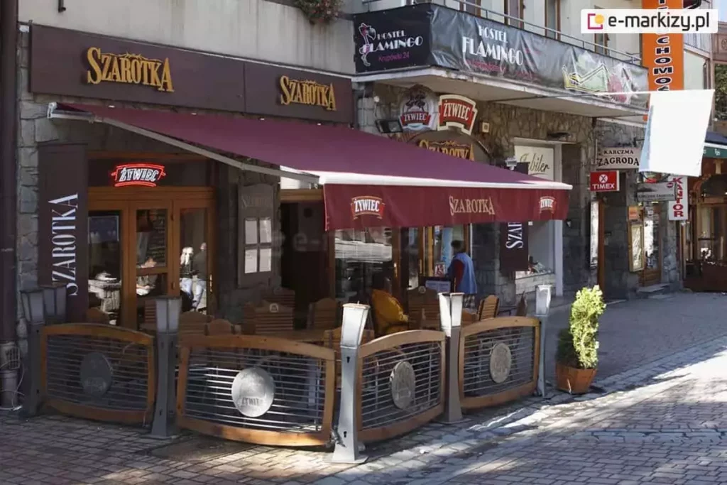 Szerokokątne ujęcie prezentuje zewnętrzny ogródek restauracyjny lokalu Szarotka, nad którym zamontowana została profesjonalna markiza reklamowa model Jamaica Aluprof w kolorze głębokiego bordowego matu. Tkanina poszycia jest napięta na solidnej, aluminiowej konstrukcji o białym kolorze, która obejmuje ramiona przegubowe wspierające wysięg markizy nad stolikami. Na przedniej falbanie markizy, zwisającej pionowo, umieszczono białe napisy reklamowe z nazwą lokalu oraz logotypami partnerów, co podkreśla komercyjne zastosowanie produktu. Markiza Jamajka skutecznie zacienia przestrzeń tarasu wyposażonego w drewniane meble ogrodowe oraz dekoracyjne ogrodzenie z elementami metalowymi i logotypami browaru. Konstrukcja markizy jest zamontowana do elewacji budynku wykonanej z kamienia i drewna, harmonizując z architekturą miejskiego deptaka. Widoczny mechanizm pozwala na duży wysięg, chroniąc klientów restauracji przed słońcem, jednocześnie pełniąc funkcję nośnika identyfikacji wizualnej marki. W tle widoczne są inne lokale handlowe i usługowe, co sytuuje produkt w przestrzeni intensywnego ruchu pieszego, gdzie markiza reklamowa Jamajka stanowi istotny element estetyczny i użytkowy fasady.