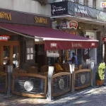 Szerokokątne ujęcie prezentuje zewnętrzny ogródek restauracyjny lokalu Szarotka, nad którym zamontowana została profesjonalna markiza reklamowa model Jamaica Aluprof w kolorze głębokiego bordowego matu. Tkanina poszycia jest napięta na solidnej, aluminiowej konstrukcji o białym kolorze, która obejmuje ramiona przegubowe wspierające wysięg markizy nad stolikami. Na przedniej falbanie markizy, zwisającej pionowo, umieszczono białe napisy reklamowe z nazwą lokalu oraz logotypami partnerów, co podkreśla komercyjne zastosowanie produktu. Markiza Jamajka skutecznie zacienia przestrzeń tarasu wyposażonego w drewniane meble ogrodowe oraz dekoracyjne ogrodzenie z elementami metalowymi i logotypami browaru. Konstrukcja markizy jest zamontowana do elewacji budynku wykonanej z kamienia i drewna, harmonizując z architekturą miejskiego deptaka. Widoczny mechanizm pozwala na duży wysięg, chroniąc klientów restauracji przed słońcem, jednocześnie pełniąc funkcję nośnika identyfikacji wizualnej marki. W tle widoczne są inne lokale handlowe i usługowe, co sytuuje produkt w przestrzeni intensywnego ruchu pieszego, gdzie markiza reklamowa Jamajka stanowi istotny element estetyczny i użytkowy fasady.