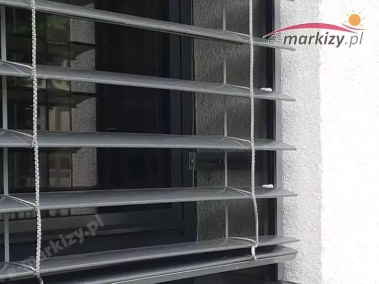 Zbliżenie na fragment zewnętrznej żaluzji fasadowej zamontowanej na oknie w elewacji budynku. Widoczne są poziome lamele wykonane z wyprofilowanego aluminium, najprawdopodobniej w kształcie litery C – z dodatkową zawiniętą krawędzią płatka lameli. Lamele są osadzone w tekstylnej drabince podnosząco-opuszczającej, prowadzonej pionowo po obu stronach żaluzji. Drabinki wykonane są z plecionych włókien technicznych – odpornych na działanie promieni UV, wilgoci i zmiennych temperatur. Okno posiada nowoczesny profil aluminiowy lub PVC z widoczną szybą zespoloną. Elewacja wykończona jest jasnym tynkiem strukturalnym. Konstrukcja żaluzji zamocowana jest we wnęce okiennej, co wskazuje na montaż podtynkowy lub we wnęce z kasetą ukrytą nad górną krawędzią okna.