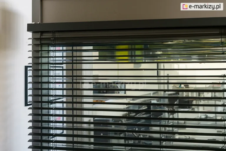 Zbliżenie na górną sekcję żaluzji poziomej C50 Slim Aluprof zamontowanej wewnątrz przeszklonego pomieszczenia biurowego lub technicznego. Zdjęcie prezentuje czarne lamele o delikatnie wygiętym profilu, które są zawieszone na systemie drabinek sznurkowych, zapewniających ich równomierne rozmieszczenie i możliwość regulacji kąta nachylenia. Górna część mechanizmu jest osłonięta solidną, ciemnobrązową maskownicą o matowym wykończeniu, która integruje się z konstrukcją ramy okiennej. Przez przezroczyste przeszklenie widoczne jest tło z elementami wyposażenia warsztatowego lub magazynowego, a po lewej stronie na białej ścianie zawieszona jest ramka z dokumentem lub certyfikatem. Detal ukazuje precyzję wykonania elementów sterujących oraz sposób montażu systemu żaluzji przy samej krawędzi górnego profilu.