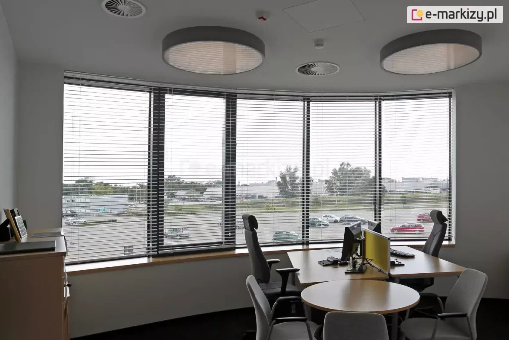 Widok z wnętrza nowoczesnego biura na panoramiczne, łukowe przeszklenie wyposażone w system wewnętrznych żaluzji poziomych C50 Slim. Srebrne, aluminiowe lamele są ustawione w pozycji otwartej, co zapewnia optymalną kontrolę nad dopływem światła dziennego przy zachowaniu widoczności na zewnętrzny parking oraz infrastrukturę miejską. W pomieszczeniu znajduje się stanowisko pracy z biurkiem o jasnym blacie, wyposażone w monitory komputerowe oddzielone żółtym panelem akustycznym oraz ergonomiczne fotele biurowe. Na suficie zamontowano dwie duże, okrągłe oprawy oświetleniowe typu plafon oraz okrągłe anemostaty systemu wentylacji. Estetyka wnętrza jest minimalistyczna, z białymi ścianami i ciemną wykładziną, co podkreśla techniczny charakter zastosowanych osłon okiennych dopasowanych do krzywizny fasady.